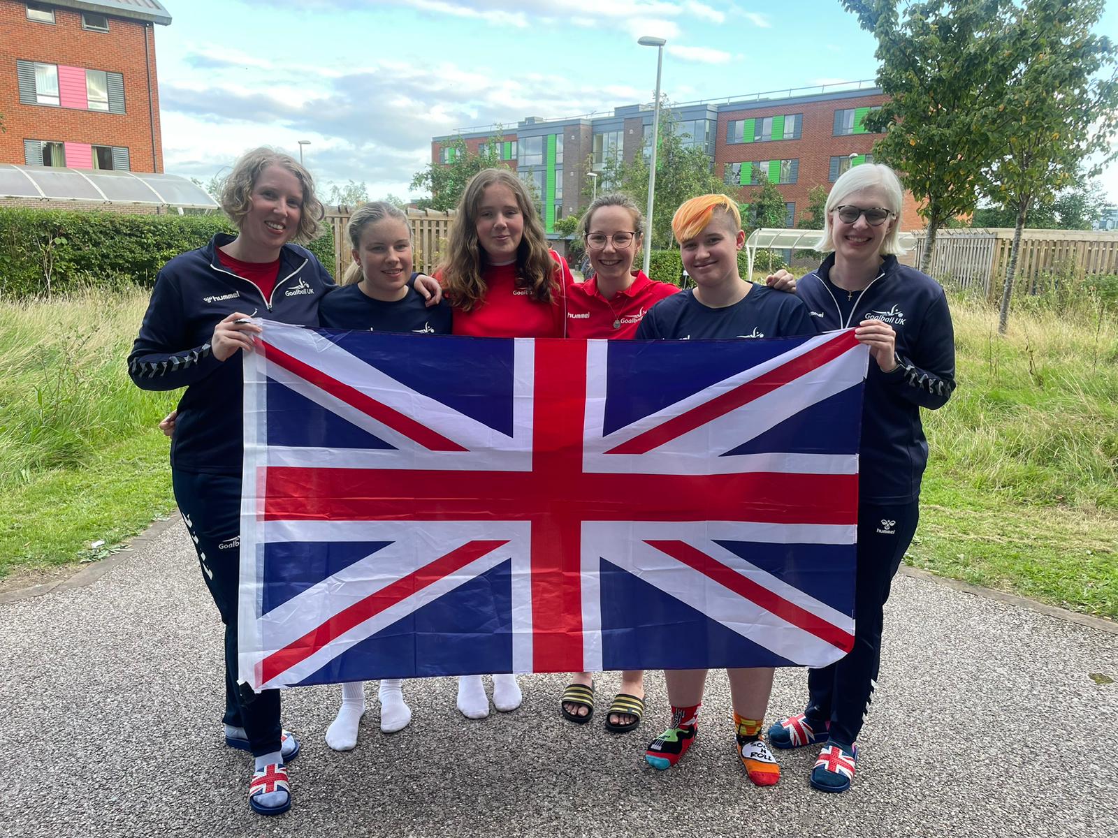 GB Women L-R Georgie, Meg, Lois, Antonia, Meme and Sarah stand outside in Goalball UK off court kit holding a Union Jack flag