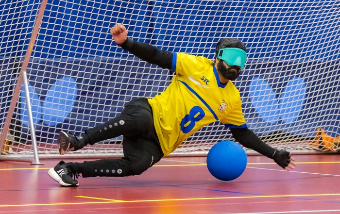 A male West Yorkshire Goalball player dives to his left to defend a goal attempt. He is wearing a yellow shirt and turquoise eyeshades