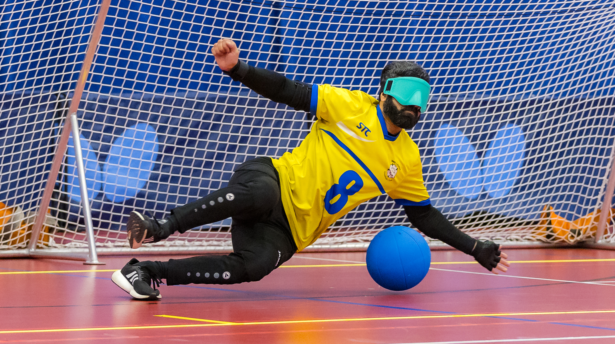 A male West Yorkshire Goalball player dives to his left to defend a goal attempt. He is wearing a yellow shirt and turquoise eyeshades