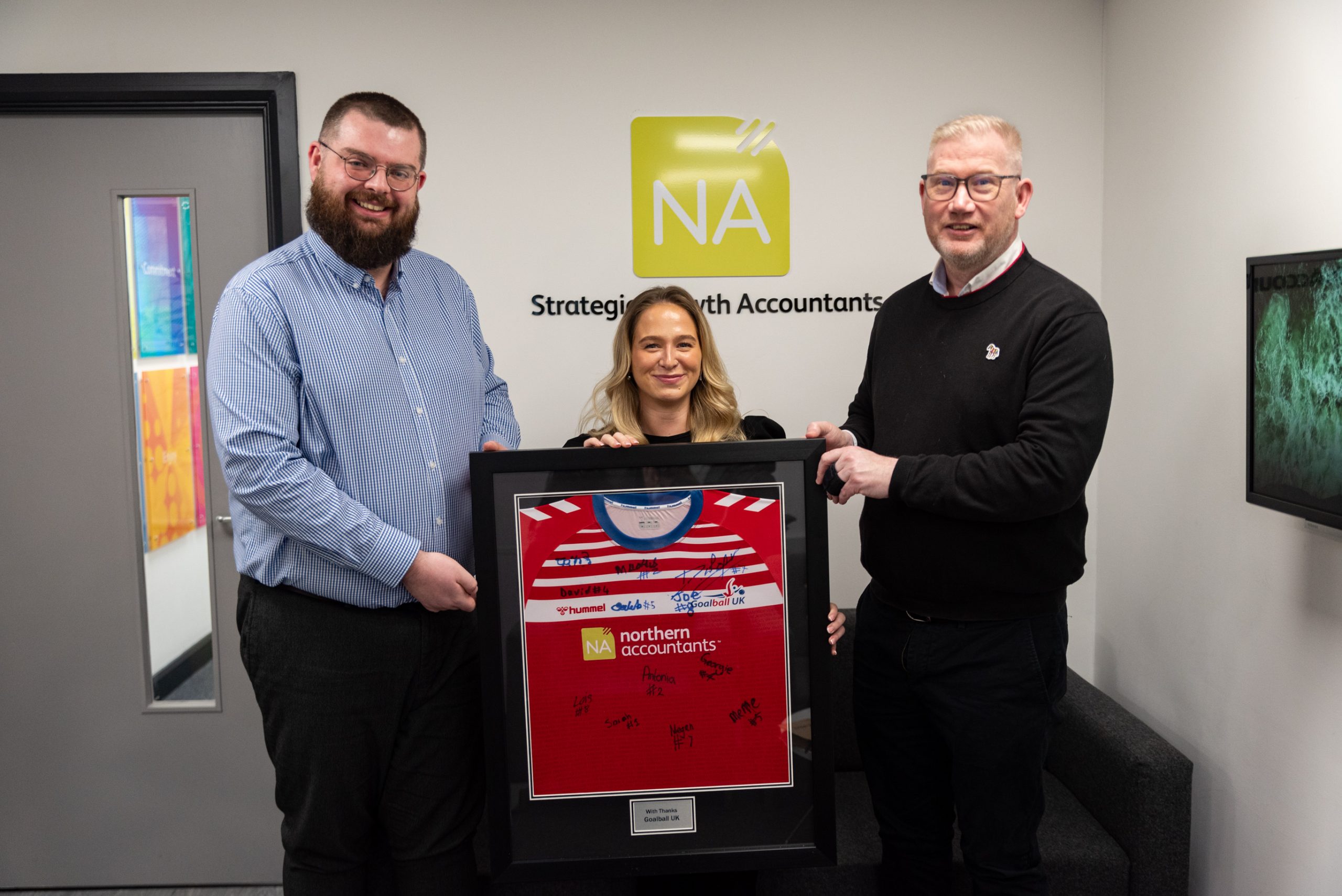 Three people in smart clothing hold up a large frame with a signed GB goalball shirt in it. Two people are from Northern Accountants and the third is Mark Winder, CEO of Goalball UK.