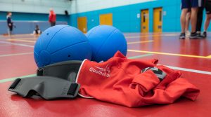 Two blue goalballs, two black eyeshades and a red Goalball UK jumper are laid on the court floor in focus, whilst the wider court is out of focus behind the items.