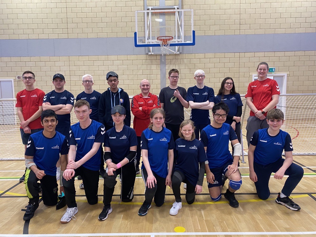 Goalball Academy group photo with players, and staff in two lines, one at the front with 7 players kneeling, and a row behind them with 9 people standing. They are in front of a goalball goal and smiling to the camera after a fun camp!