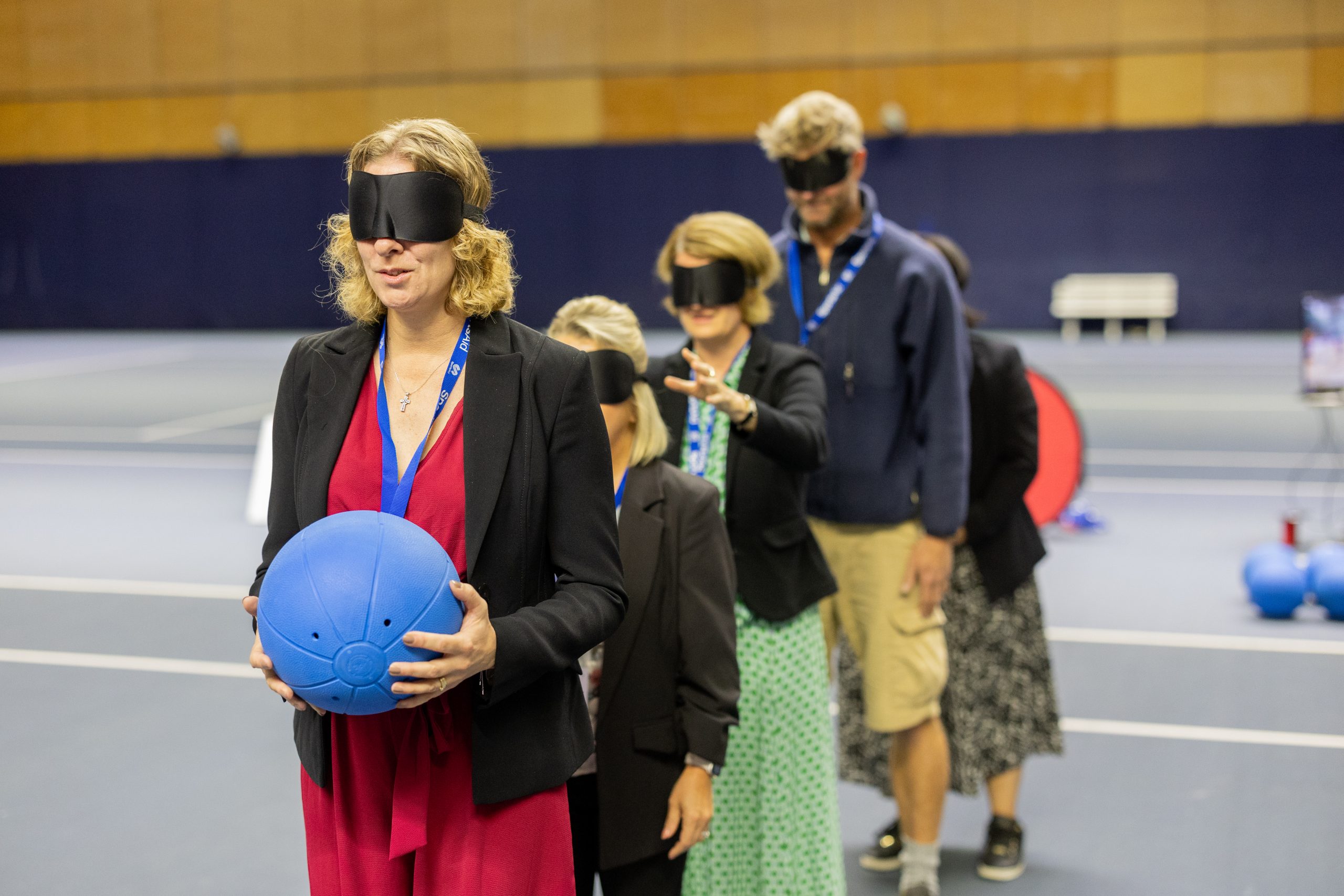 Men and women in smart corporate clothes stand in a line. They are wearing black eye shades and the woman at the front holds a goalball ready to play a game of 'over and under'