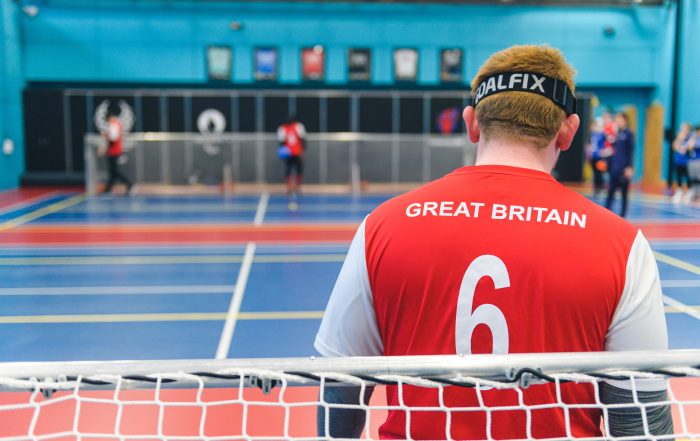 Photo taken from behind a goalball goal with Chris Colbert looking towards the other end of the court, wearing a red GB shirt.