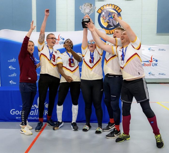 Coach Faye Dale, Matt Loftus, Caleb Nanevie, Lois Turner, Stuart Hudson, and Peter Doyle of Northern Allstars in their white jerseys, all with a hand on the Goalfix Cup Trophy which they're lifting in the air in celebration!