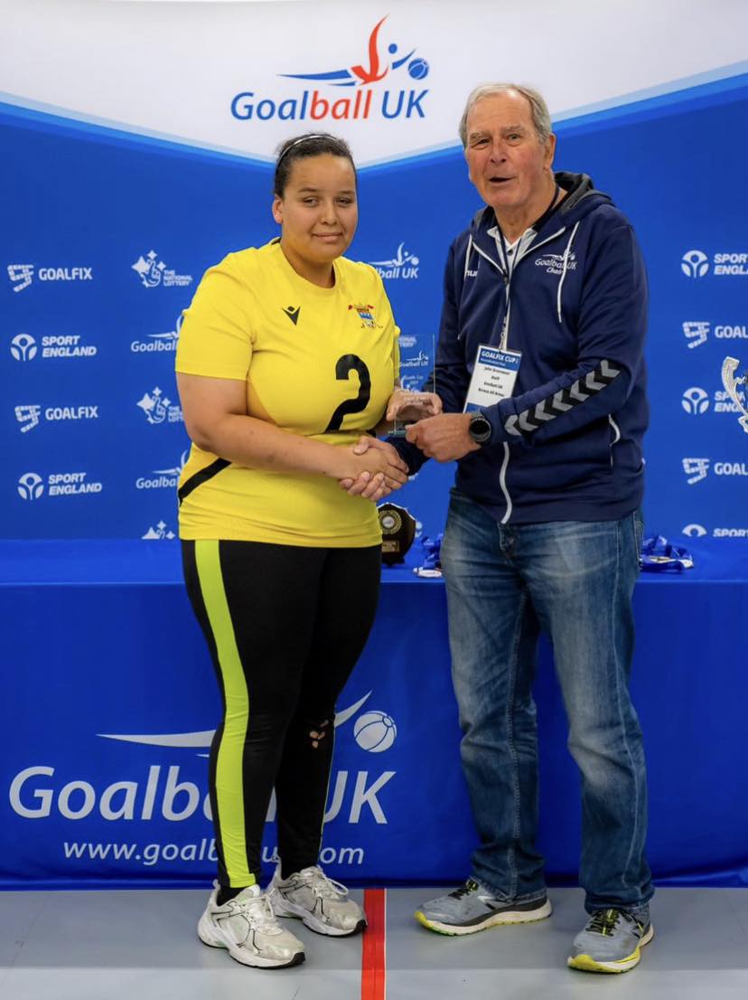 Chelsea Hudson stands with her award and poses for a photo against a Goalball UK branded banner