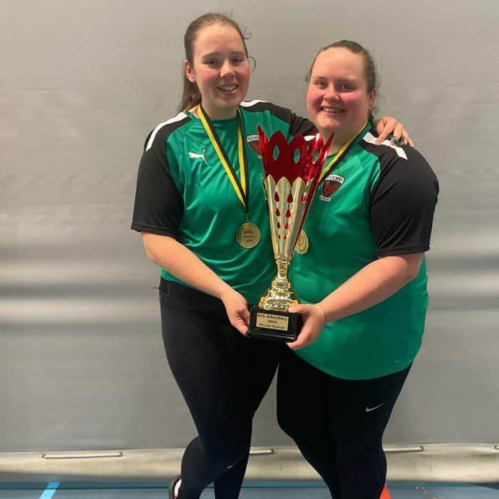 Lois Turner and Kali Holder in their green Fucshe Berline jerseys. They are stood together, each holding the European Champions League Trophy. They're stood in front of a white wall.
