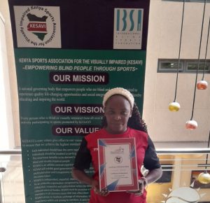 Florence stood with her goalball leaders certificate whilst wearing a red Goalball UK shirt, in front of a Kenya Sports Association for the Blind banner.