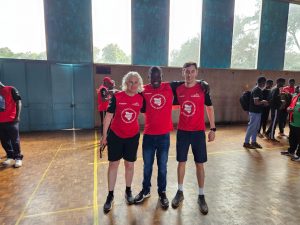 Kathryn, Stephen, and Alex stood together in a hall, all smiling to the camera. There are people standing in the background of their photo.
