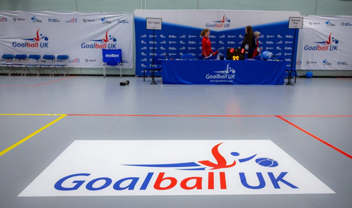 5R0A2723 Image shows the grey court floor of the Goalfix Cup 2024 with the Goalball UK logo overlayed onto it. There are court lines on show in the neutral area of the court. In the background there is the officials table.