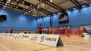 Wide angled shot of the court with sponsor banners down one side and a black screen with Goalball UK in white text above