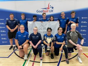 Goalball Academy players across two rows, one standing, one kneeling. The row kneeling are proudly holding the newly won Trophy by GB Women. Row standing behind are holding their Goalball Academy flag.