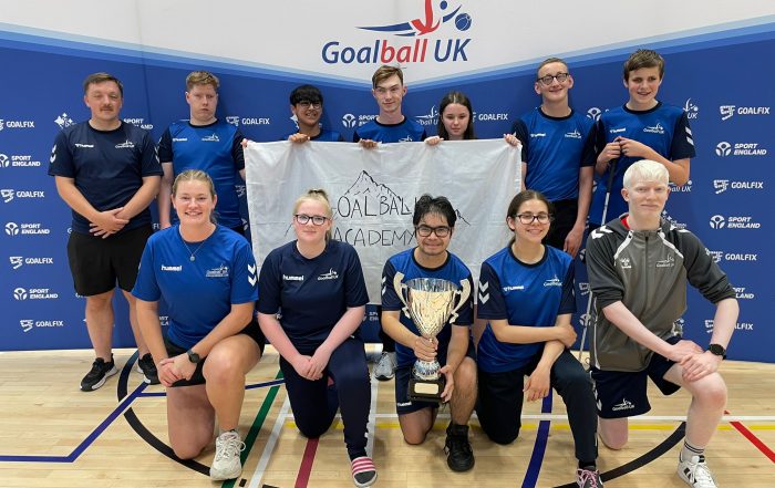 Goalball Academy players across two rows, one standing, one kneeling. The row kneeling are proudly holding the newly won Trophy by GB Women. Row standing behind are holding their Goalball Academy flag.