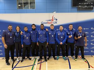 Brazil (U23) athletes and coaches pose for a group photo in their blue kit in front of the Goalball UK banner