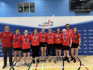 Team Canada group photo in their red kit stood with their coaches in front of a Goalball UK branded banner