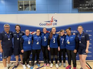 GB Women group photo in their kit stood with their coaches infront of a Goalball UK branded banner