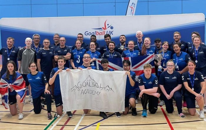 GB Women, GB Men, and the Goalball Academy players and staff in one big Performance Pathway photo. Everyone is huddled together with their arms around one another after a successful competition and camp. Players at the front are holding the Goalball Academy flag.