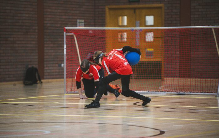 A female player for South Wales about to throw a goalball during a game, wearing a red jersey with black leggings. She is taking the shot from the centre position.