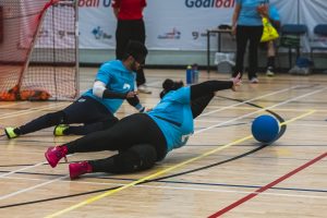 A female player for Birmingham Goalball Club diving to save a shot during a game. She is wearing a light blue jersey and is playing in the centre position.