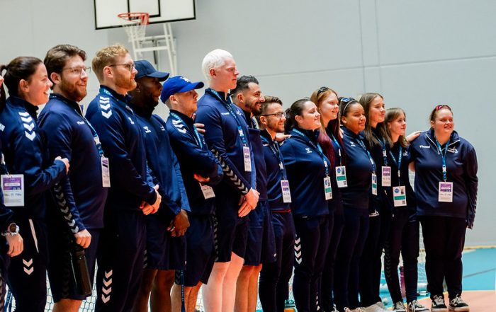 Great Britain Men and Women standing together in a line, side by side, at the opening ceremony of the IBSA goalball European A Championships. Everyone is wearing navy blue Great Britain jackets, tracksuit bottoms or shorts.