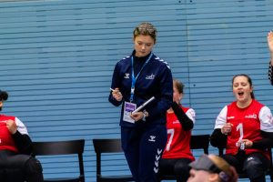 Ella Strachan, Women's assistant coach standing in the team bench area making a note in her book during a game. Behind her right shoulder sat down is player Emma Tammarro who is shouting in celebration!