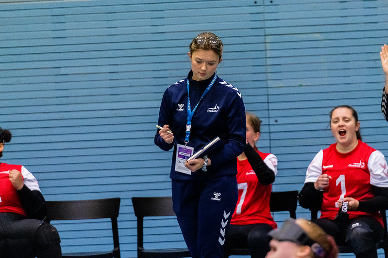 Ella Strachan, Women's assistant coach standing in the team bench area making a note in her book during a game. Behind her right shoulder sat down is player Emma Tammarro who is shouting in celebration!