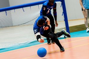Dan Roper during a game for Great Britain in his blue shirt, releasing a goalball. His left leg is planted on the floor, right leg is behind it in the air at knee height. His head is facing away as he gives maximum effort to throw the ball with his right arm!