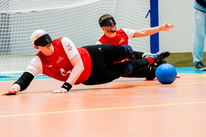 David Scott and Joe Roper during a goalball game for Great Britain, they're both on the floor just about to dive for a goalball which is coming towards them. They're wearing their red GB shirts.
