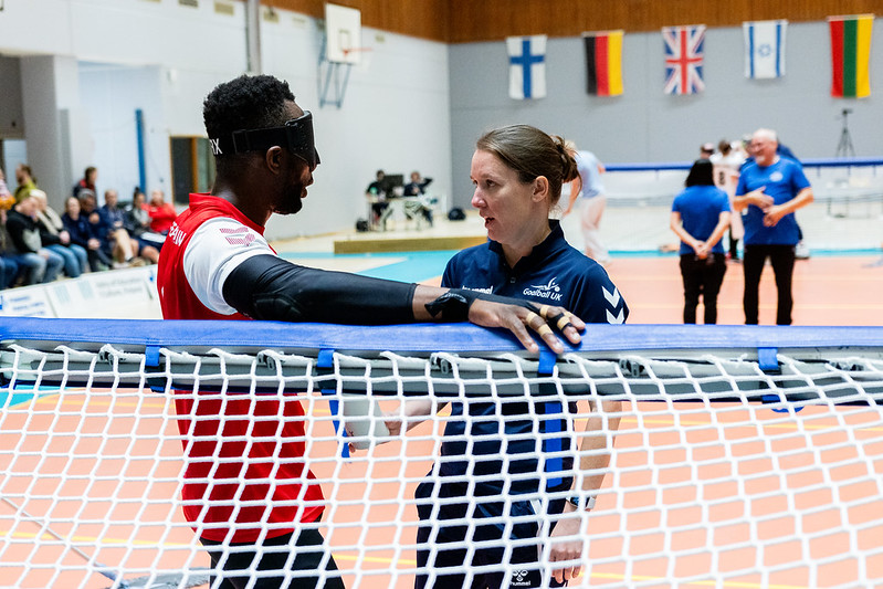 Head Coach Faye Dale-Cook speaking to Caleb Nanevie at back centre during a timeout before a penalty. Faye is giving Caleb some advice before the penalty is taken. There are two referees in the background in the middle of the court.