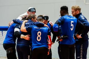 GB Men's team in a huddle with arms around one another at the end of the IBSA Goalball European A Championships. They're all in their blue GB shirts, while coaches Faye Dale-Cook and Alex Jones are speaking to them.