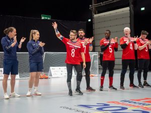 GB Men's team lineup along the high ball line while the teams are announced. Joe Roper is standing in front of everyone as his name is called out, he has his arm up in the air. Faye Dale-Cook is on the left of the image clapping to cheer Joe on!