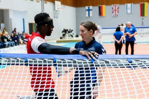 Faye speaking to player Caleb Nanevie at a timeout during a penalty midgame. They are at back centre of the court, with the two referees visible in the background.