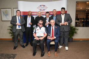 A group photo of all the award winners in front of a British Blind Sport banner. 6 people are stood with another 2 people seated. They are all smartly dressed and holding their awards. 