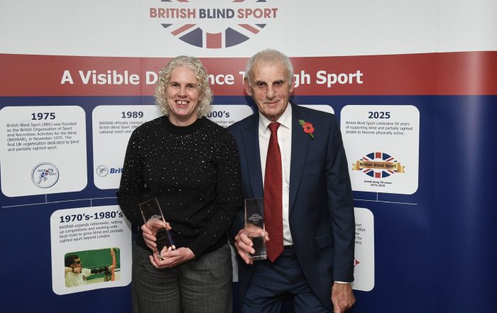 Kathryn and Clive, stood in front of a British Blind Sport banner, proudly holding their awards which are made out of glass and engraved with their names.