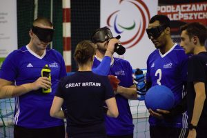 Coaches Faye, and Alex Bunney speak to players during a timeout at a previous competition. Matt Loftus, Dan Roper, and Filmon Eyassu are on court taking on fluids whilst listening to Faye and Alex.