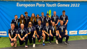 The Great Britain Men and Women's squad at the European Para Youth Games 2022. Players and staff are together for a group photo with their bronze medals, across two rows. Everyone is wearing navy blue tracksuits and are in front of a bright blue banner.