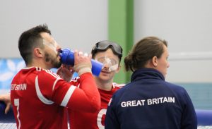 Coach Faye Dale-Cook, and players Dan Roper and Michael Sharkey standing together near a goalball goal during a competition.