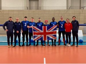 A GB team photo ahead of a previous European A Championships. All players are in their blue GB jerseys, and players and staff are standing together along a goalball goal.