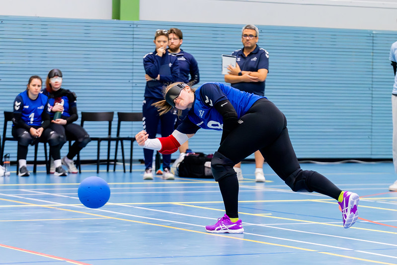54828145729_cb52d012dd_c Lois Turner playing for Great Britain at the IBSA European A Championships. Lois is lunging to throw the ball with her right arm. In the background, coaches Ella and Gary watch on.