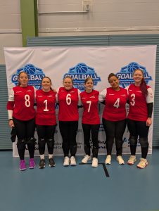 GB Women's squad at the IBSA European A Championships. Team photo, of all players in their red and white jerseys. All six players are standing with their arms around one another and smiling.