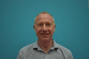 Peter Elliott standing in front of a turquoise wall in a smart buttoned, striped shirt. He is smiling to the camera.