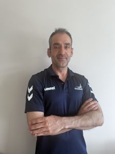 Ferhat Esatbeyoglu profile photo, Ferhat is standing in front of a white painted wall in a navy blue Goalball UK shirt. He has his arms folded and is smiling to the camera.