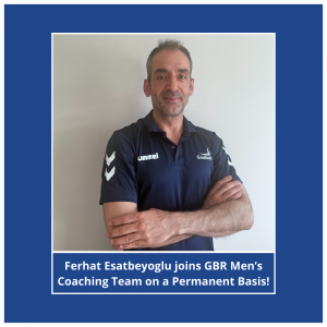 Ferhat Esatbeyoglu profile photo, Ferhat is standing in front of a white painted wall in a navy blue Goalball UK shirt. He has his arms folded and is smiling to the camera. In the background was a blue background, with a white border.