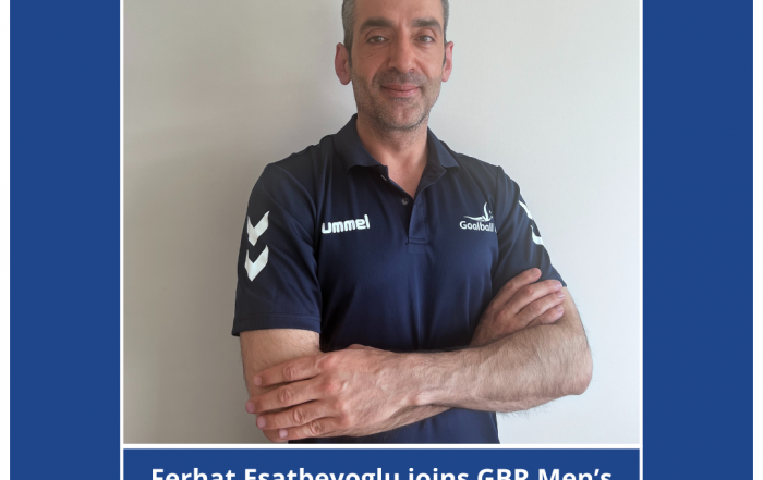 Ferhat Esatbeyoglu profile photo, Ferhat is standing in front of a white painted wall in a navy blue Goalball UK shirt. He has his arms folded and is smiling to the camera. In the background was a blue background, with a white border.