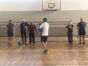 A group of players stand in a sports hall near a goal net, some holding goalballs. Alex Bunney in the center, wearing a "Great Britain" shirt, instructing the group.