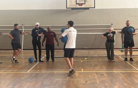 A group of players stand in a sports hall near a goal net, some holding goalballs. Alex Bunney in the center, wearing a "Great Britain" shirt, instructing the group.