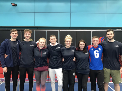 Photograph of a group of eight people standing side by side indoors against a blue wall with black lockers behind them. Most individuals wear matching black shirts with a small white logo, while Alex Cockerham second in from the right wears a blue sports jersey with the number 6