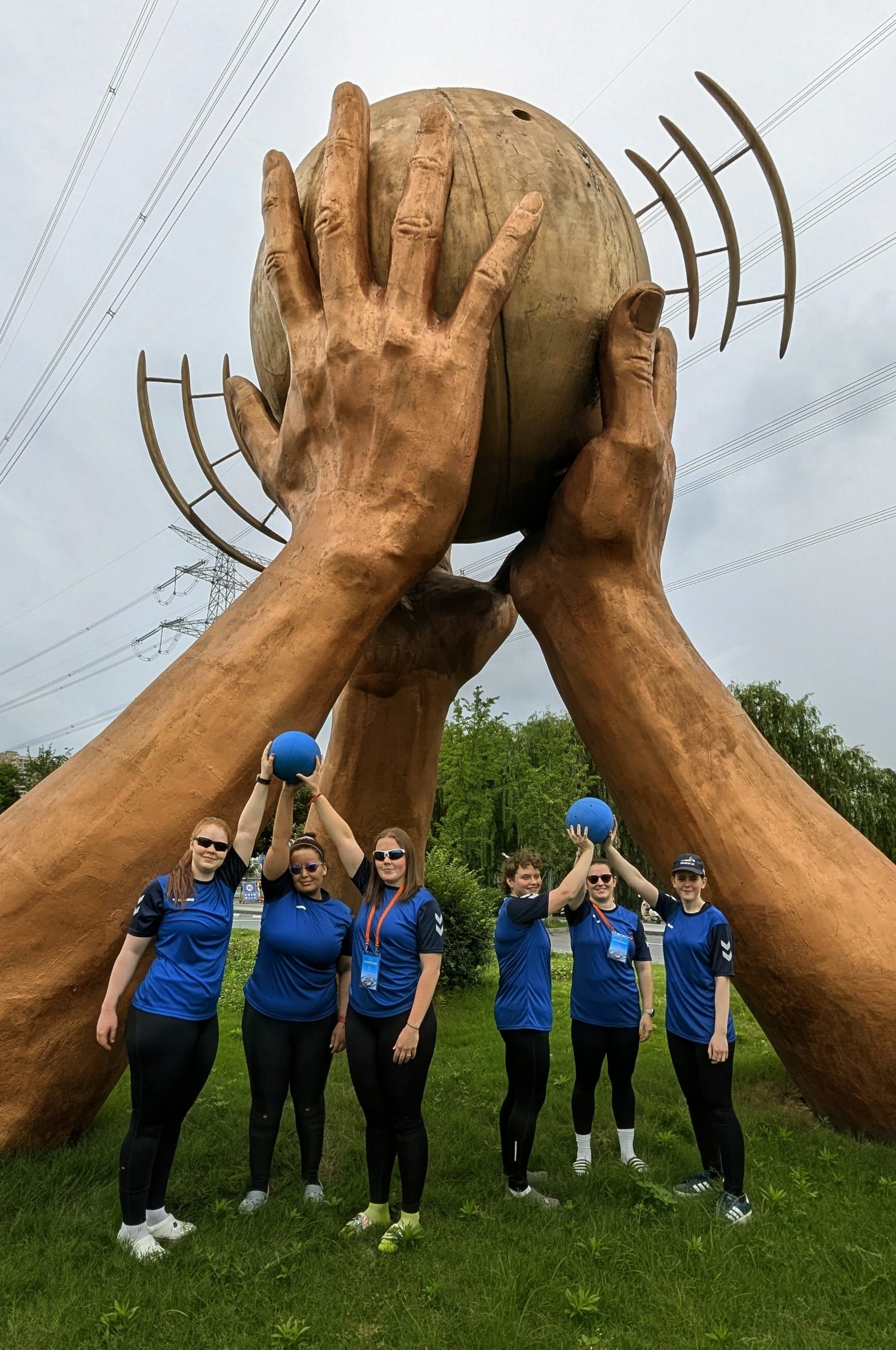 The GB Women's Squad wearing their blue shirts and holding goalballs are standing in front of a large sculpture of two hands holding a goalball up in the air. The goalball sculpture is approximately 3 times taller than the players.