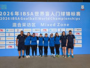 Group photo of the GB Women's squad and staff, stood in front of a large IBSA Goalball World Championships board.