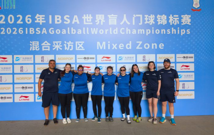 Group photo of the GB Women's squad and staff, stood in front of a large IBSA Goalball World Championships board.